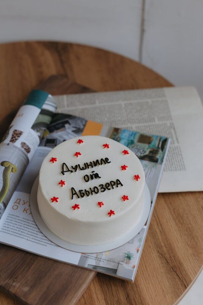 White cake with Cyrillic text, placed on open magazines, wooden table background.