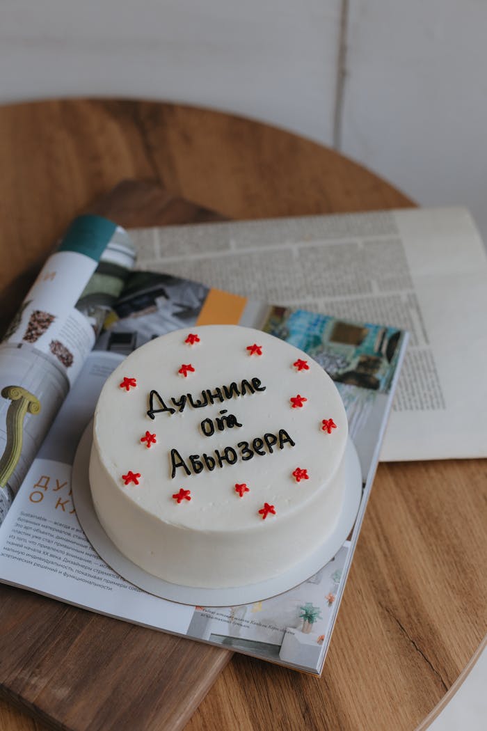 White cake with Cyrillic text, placed on open magazines, wooden table background.
